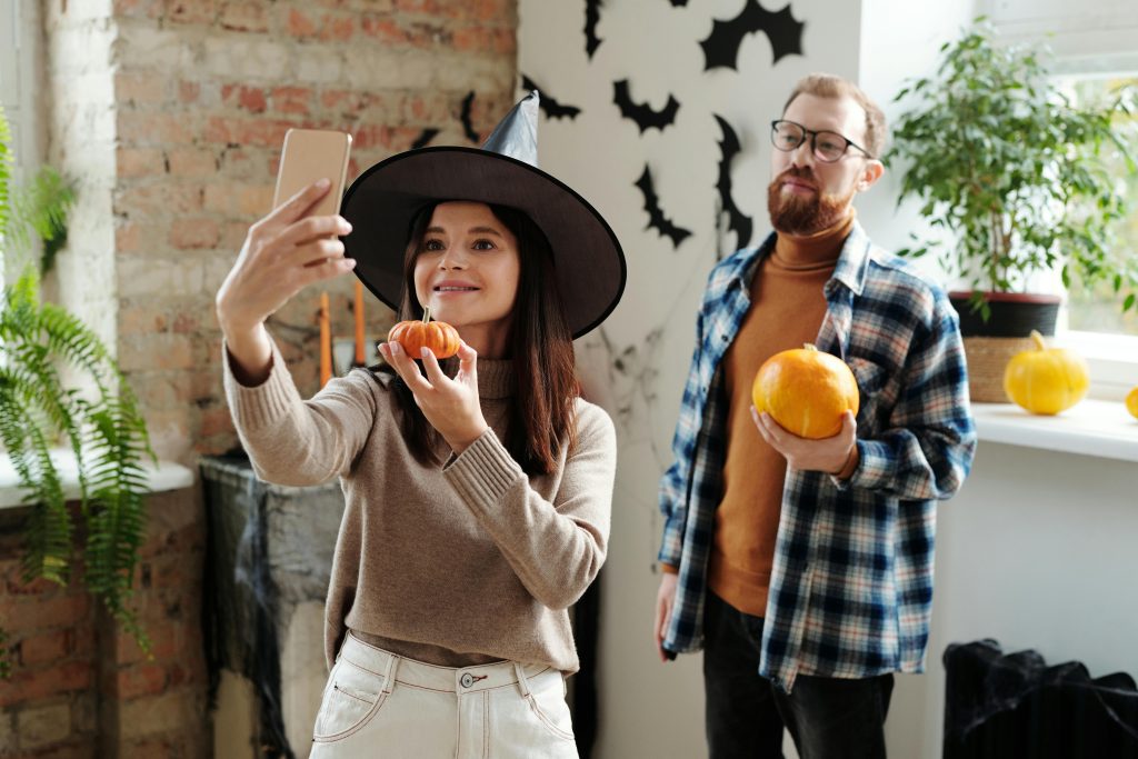 Woman in witch hat taking a selfie with a small pumpkin, with a man holding a pumpkin in the background, all set in a halloween-decorated room.