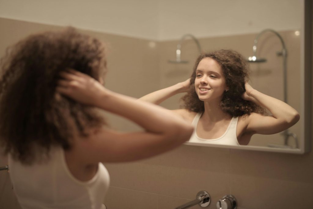 A young woman with curly hair smiles at her reflection in the bathroom mirror, capturing a moment of self-assessment and the complexities of self-esteem.