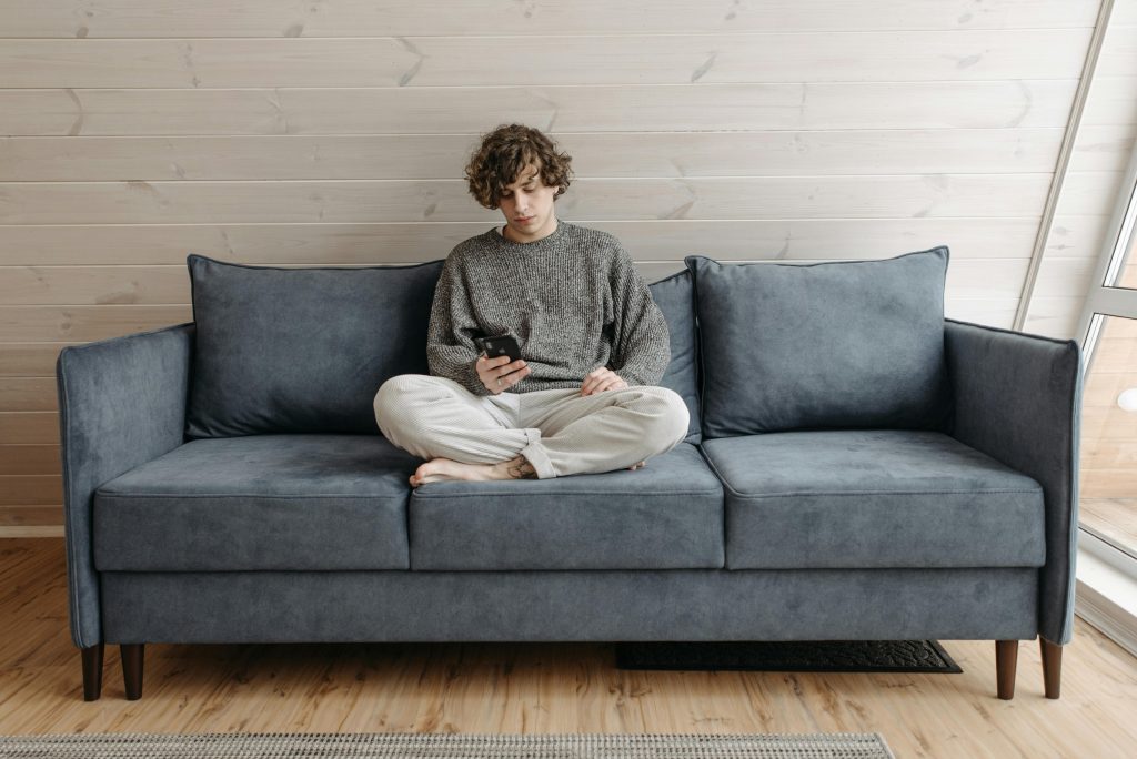 A young man sitting cross-legged on a blue couch, looking contemplative while using his smartphone, possibly reflecting on existential questions.