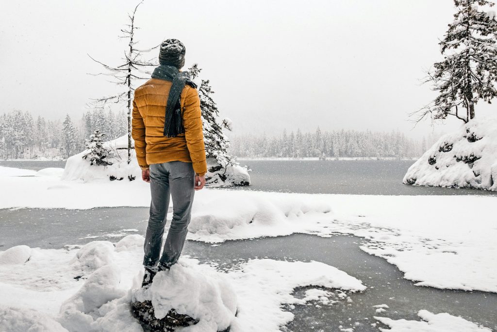 Person in a yellow jacket standing by a frozen lake surrounded by snow-covered trees, depicting the winter blues.