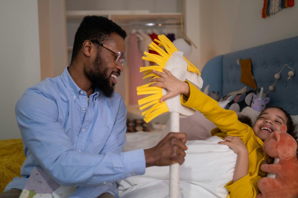 A father and his daughter enjoying playful interaction in a bedroom, the child dressed in a bright yellow shirt, engaging with a creative plush toy, illustrating a lively and nurturing family environment.