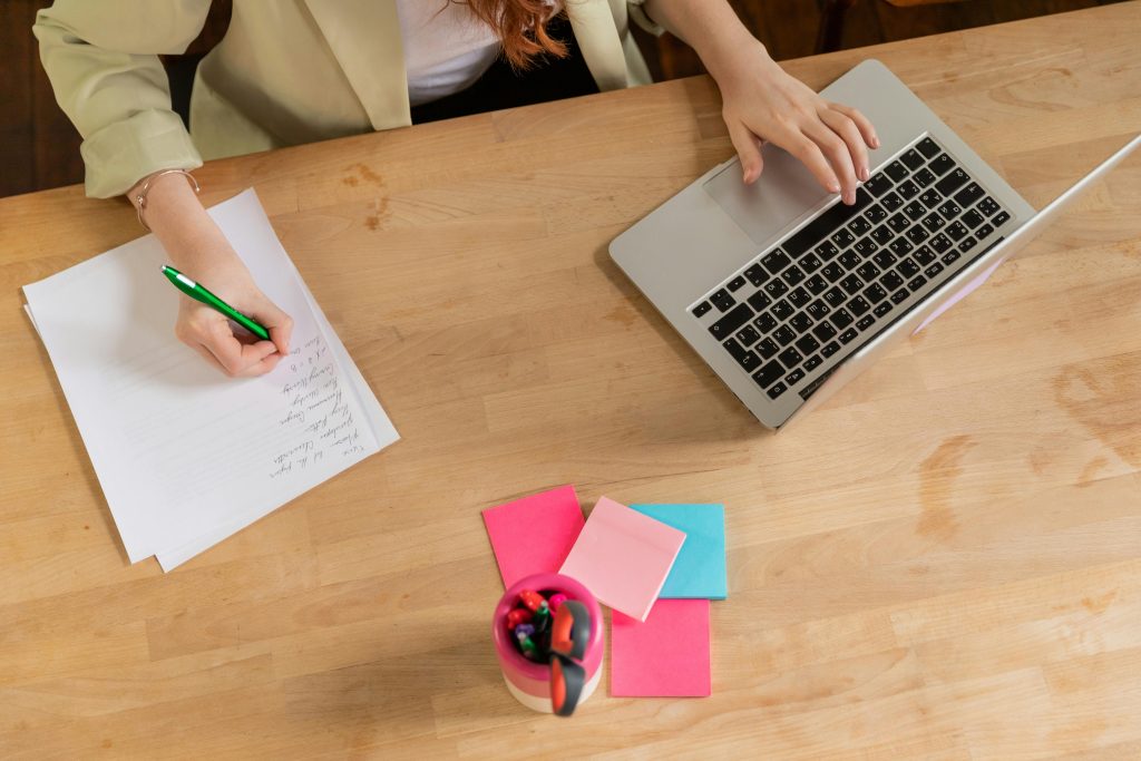 A workspace scene with a woman writing on a paper next to a laptop, sticky notes, and writing supplies, depicting organizational strategies to manage executive dysfunction.