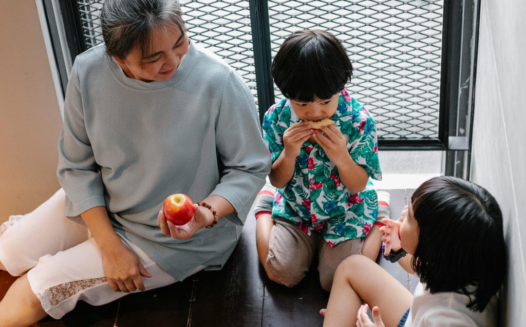 An older woman and two young children sitting on a balcony, sharing a snack and a moment of connection, symbolizing generational trauma healing in a family setting.