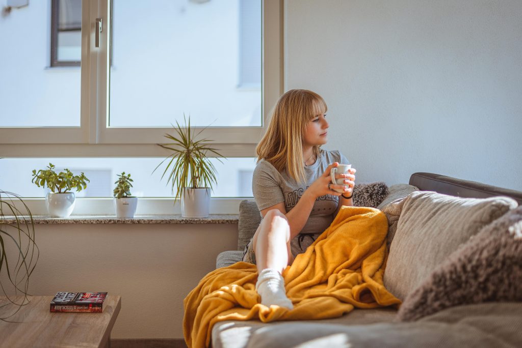 A woman relaxing on a couch, covered with a cozy blanket, holding a cup, and looking out the window, embodying a peaceful moment of self-care and love.
