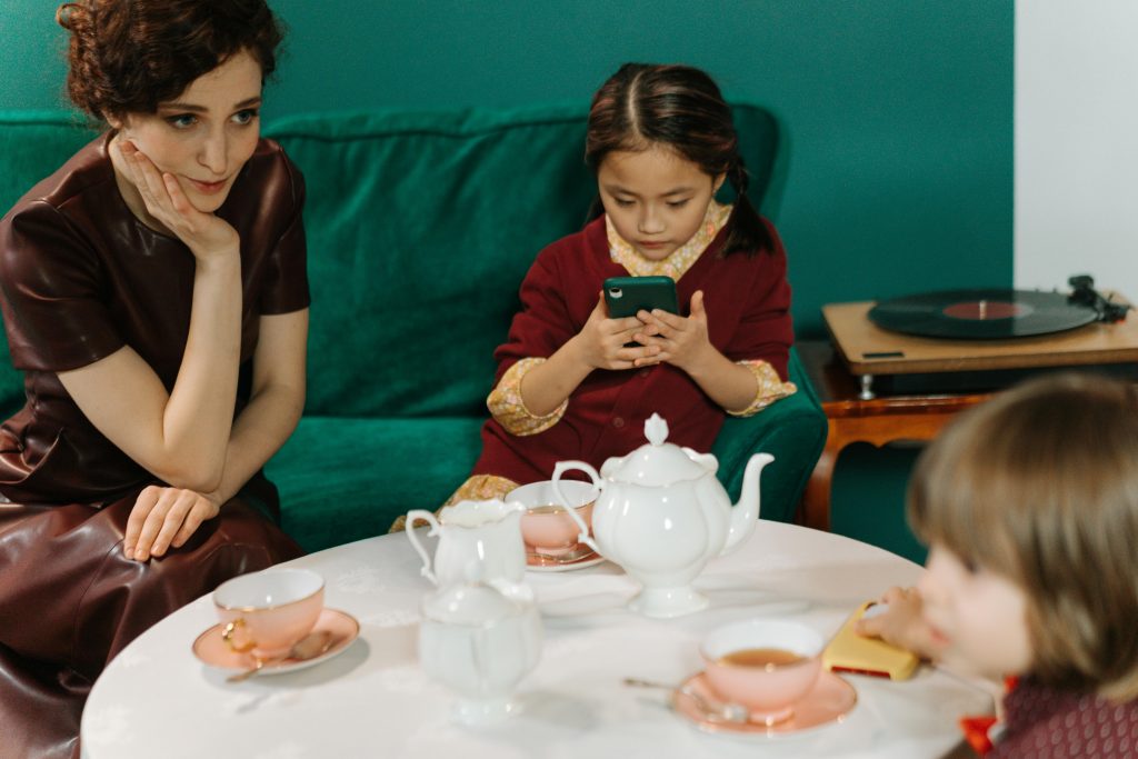 A mother observing her young children during tea time, capturing a moment of neurodivergent behaviour as one child is engrossed in a smartphone.