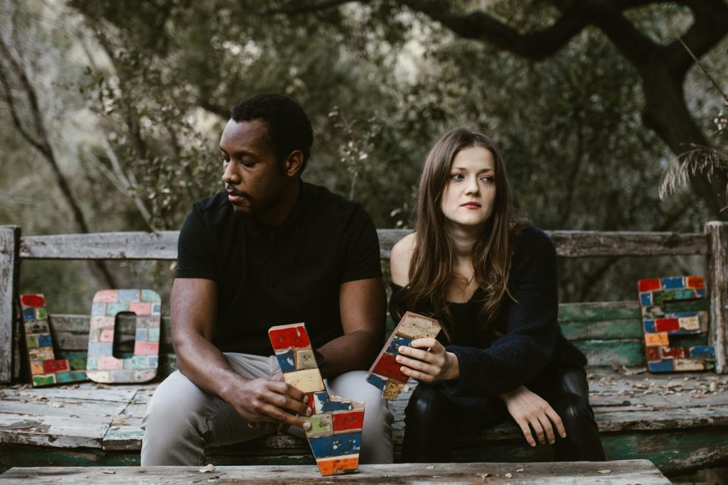 A couple sits distantly on a rustic park bench, each holding blocks of memories or grievances, symbolizing the building tensions in a toxic relationship, amidst a serene wooded backdrop.