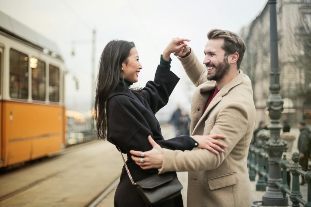 A couple playfully dancing on a city street with a tram passing by in the background, their joyous expressions belying the underlying complexities of a toxic relationship.