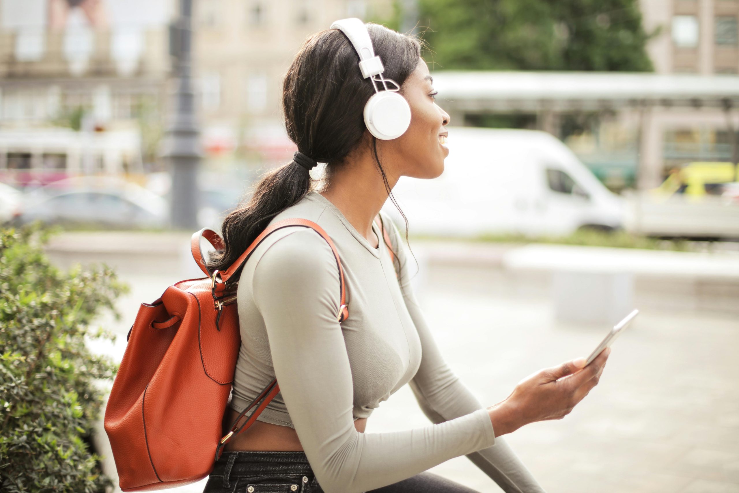 Woman-Listening-To-Music-With-Headphones A young woman sitting outdoors with headphones on, smiling while holding her phone, symbolizing relaxation and self-care.
