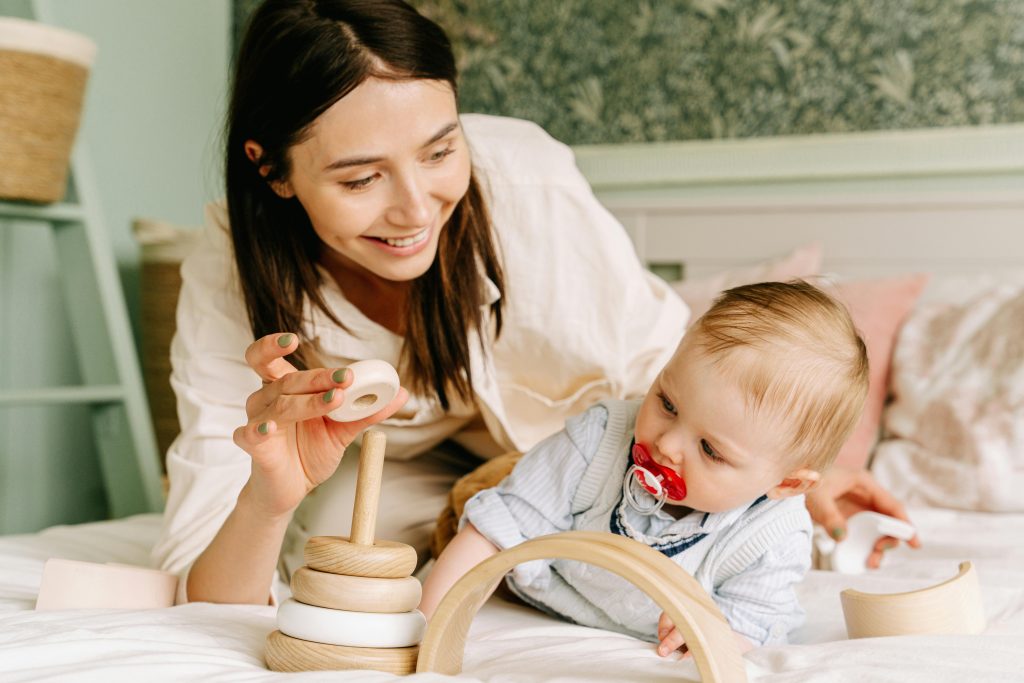 A smiling young mother playing with her baby on a bed, engaging with a wooden toy, representing supportive and interactive elements in postpartum depression treatment.