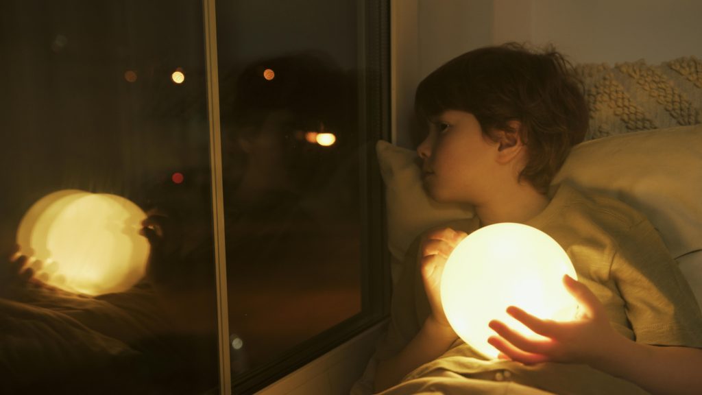 A young child gazes out the window while holding a glowing orb, symbolizing the introspection and emotional withdrawal associated with the short-term effects of domestic violence.