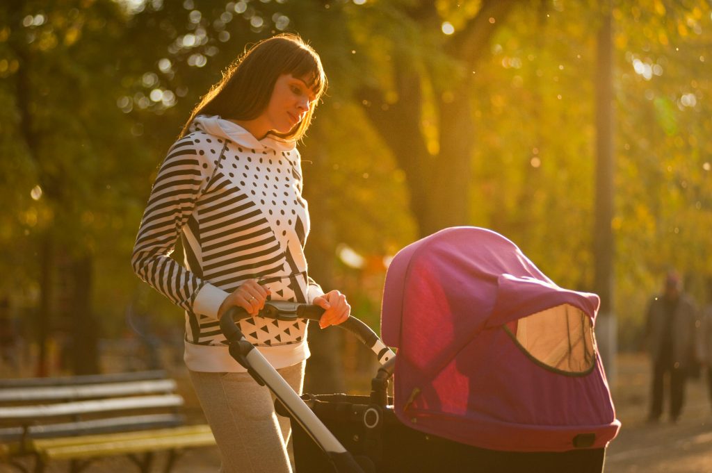A mother walks with her baby stroller in a park during autumn, appearing contemplative and distant, which may indicate symptoms of postpartum depression.