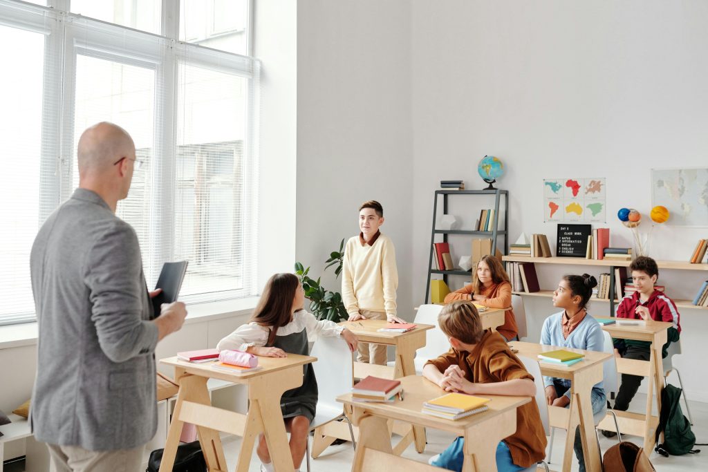 A diverse group of students in a bright classroom listen intently as a teacher discusses, embodying the theme 'what it means to be nervous' during a school presentation, highlighting the interaction and attentive expressions.