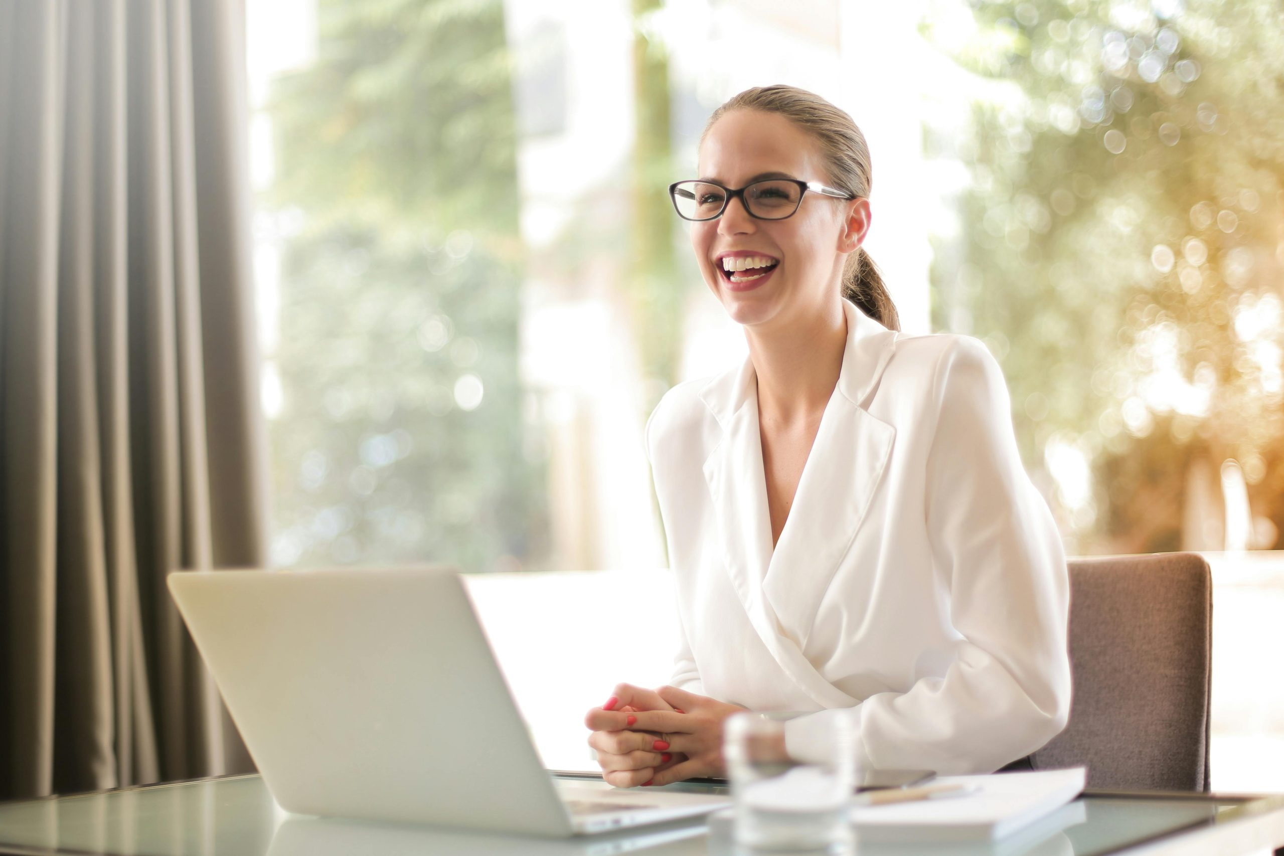 Smiling woman in a white blazer at her laptop, representing empowerment, clarity, and renewed confidence after trauma recovery therapy.