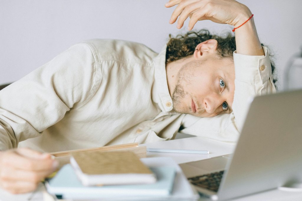 A tired man rests his head on a desk beside a laptop and notebooks, showing signs of burnout and mental exhaustion.