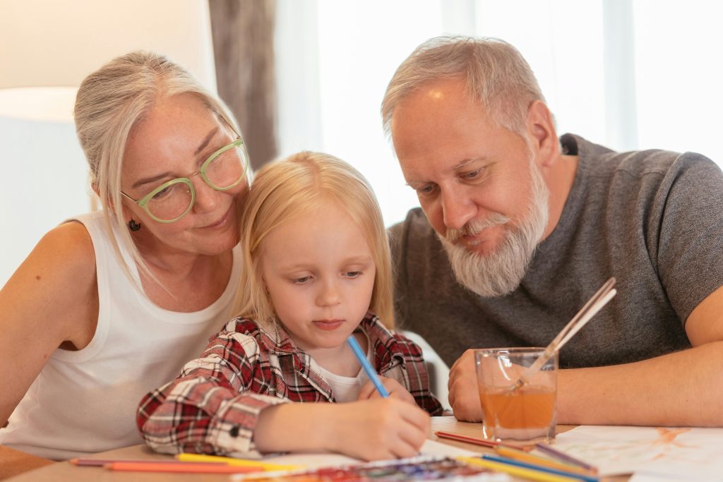 Grandparents lovingly support a child as she draws, symbolizing healing, support, and the breaking of intergenerational trauma cycles through nurturing presence and creativity