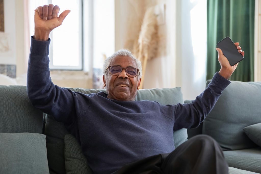 An older man sits comfortably on a couch, smiling and raising his arms in a joyful gesture, symbolizing emotional resilience and recovery from trauma's impact on the brain.
