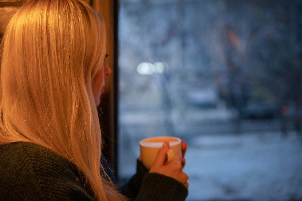 A woman with long blonde hair holds a mug and stares out of a window on a cold, gloomy day, symbolising emotional withdrawal and the mental effects of trauma on the brain.