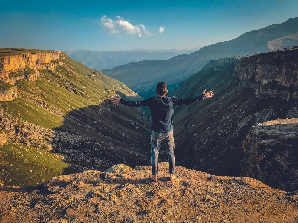 Man standing barefoot on a cliff with arms wide open, facing a vast mountain valley.