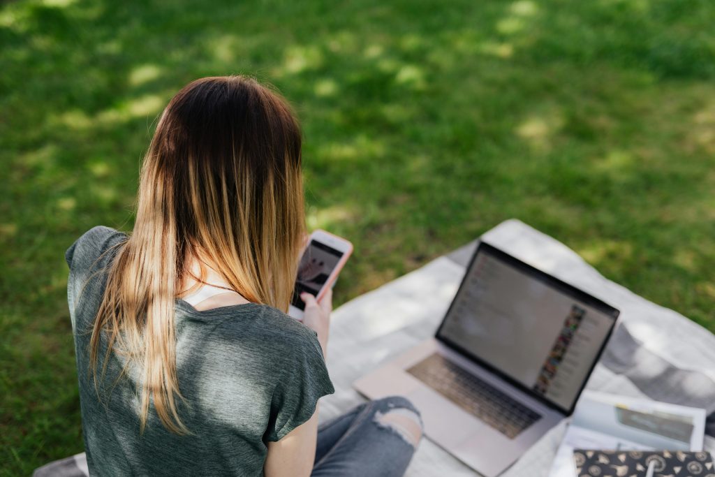 Teen girl scrolling on phone beside an open laptop on a blanket outside.