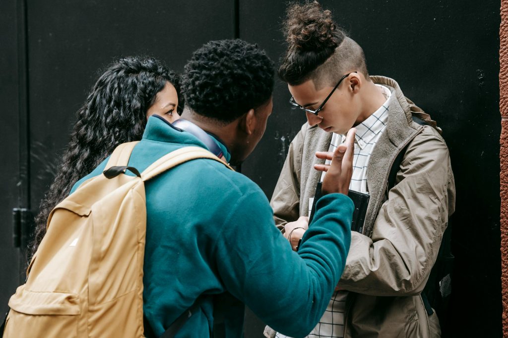A young man confronts another who is looking down and defensive, while a third person watches closely, demonstrating interpersonal conflict or aggression.