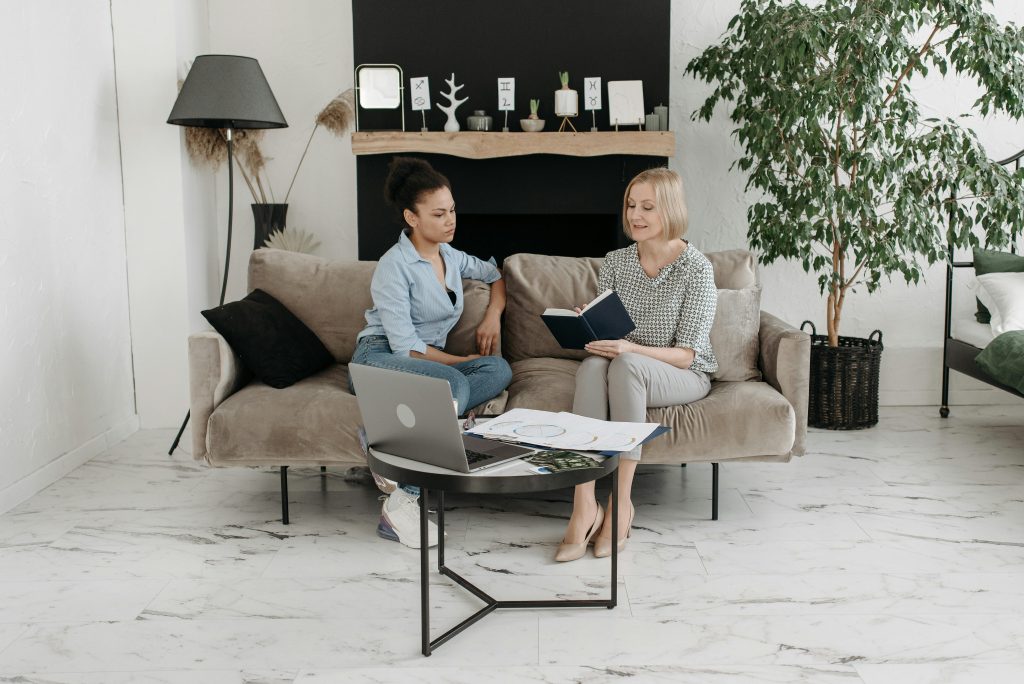 Two women sitting on a sofa with a laptop and notebook, engaged in a focused therapy session about treatment options.