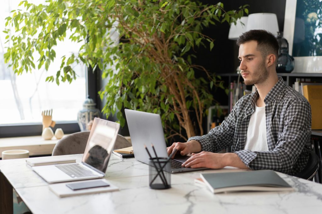 A young man sitting at a desk with two laptops, appearing focused but emotionally drained, reflecting the theme of being exhausted yet still functioning.