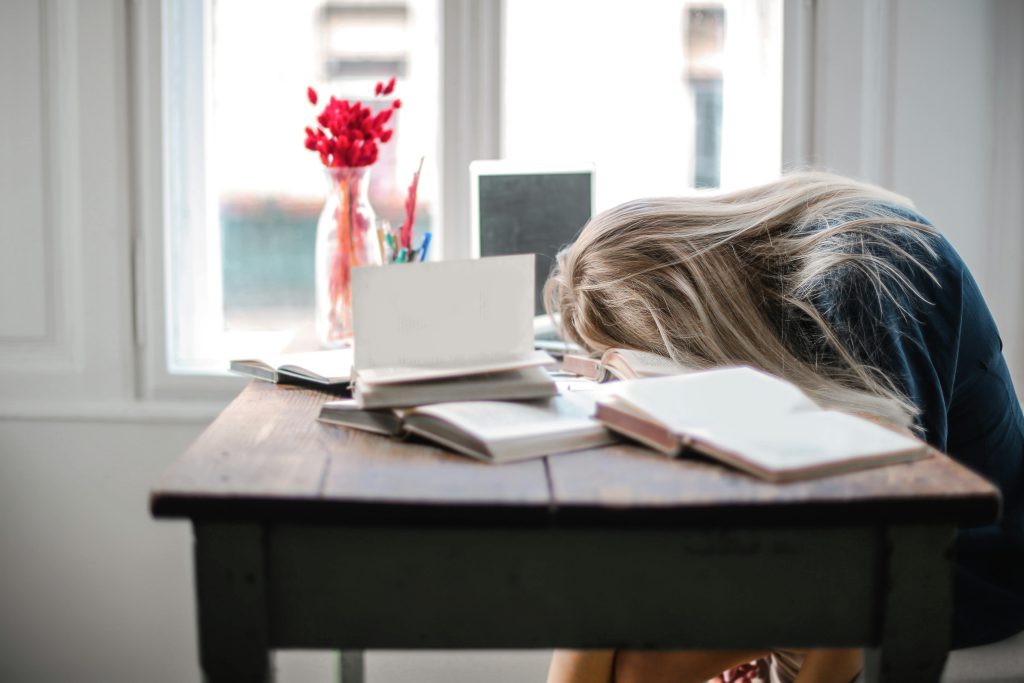 Overwhelmed woman resting her head on a desk cluttered with open books, reflecting mental exhaustion and work-related burnout.