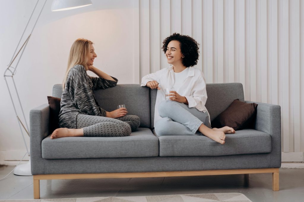 Two women smiling and chatting on a grey sofa, representing integrative therapy in a relaxed setting.