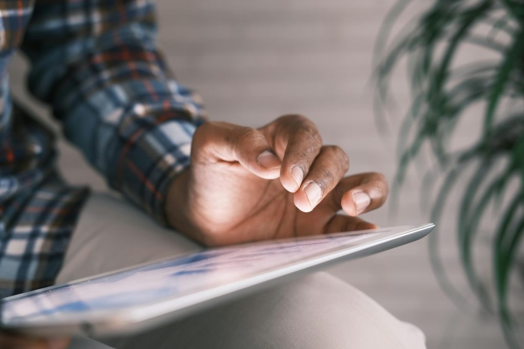 Close-up of a person using a tablet device, wearing a plaid shirt and sitting indoors, with a leafy plant blurred in the background