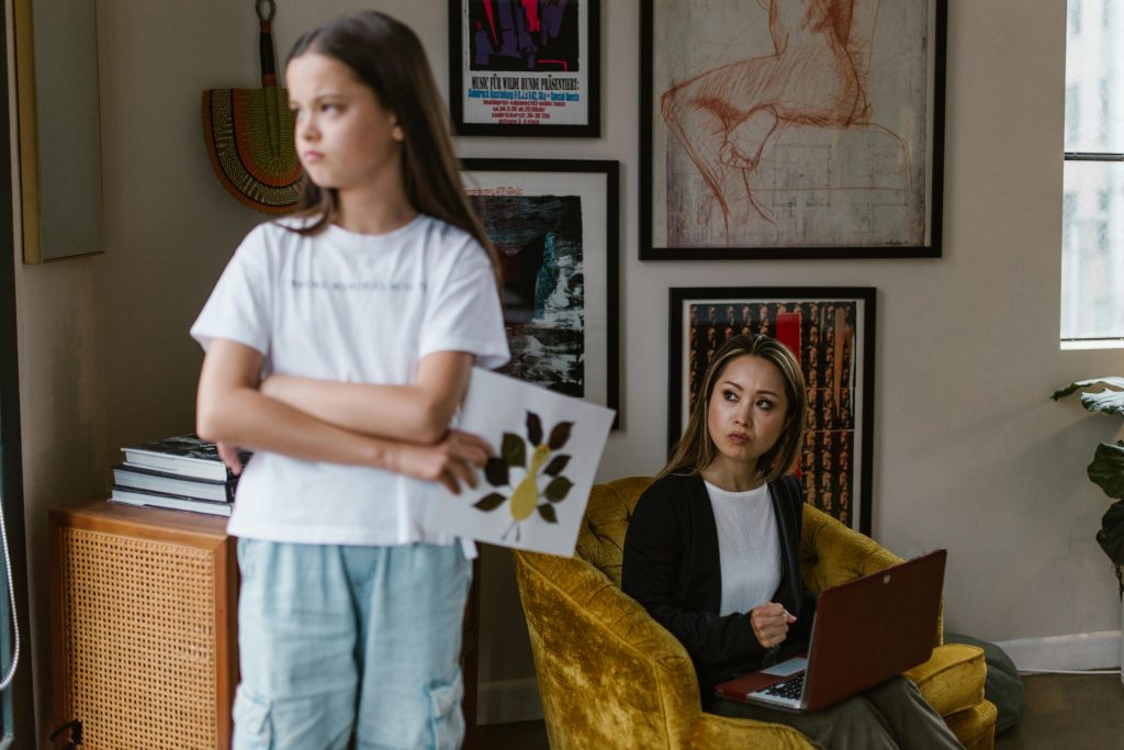 Young girl standing with arms crossed while mother looks at her from chair with laptop