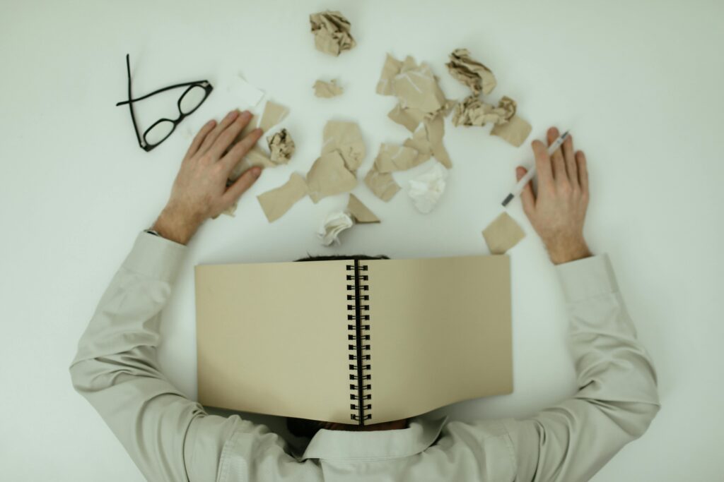 Person with notebook over head and crumpled papers on desk, showing why do i feel overwhelmed all the time