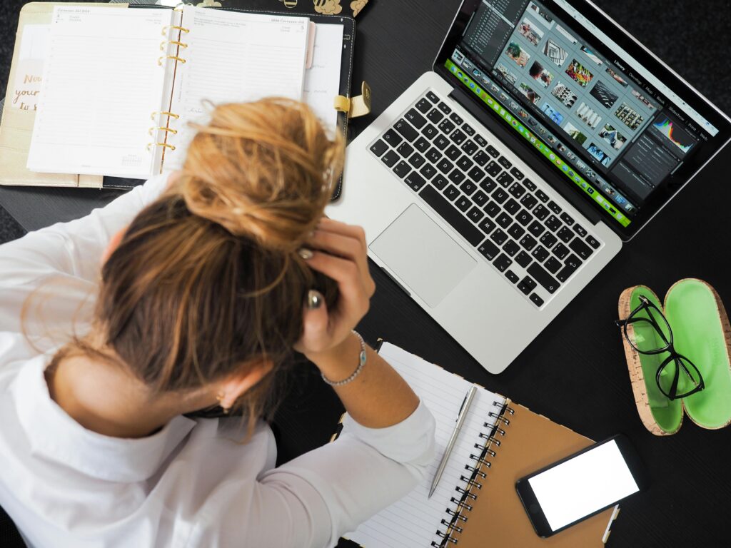 Person holding head at cluttered desk with laptop and planner, showing why do i feel overwhelmed all the time